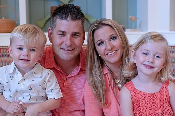Two adults and two young children sit closely together and smile at the camera.