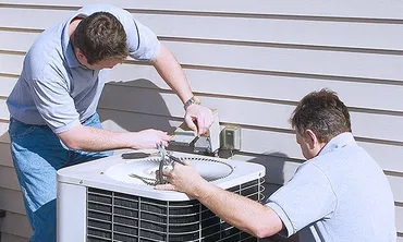 Two men work on the open top of an outdoor air-conditioning unit beside a wall.