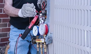 A person wearing gloves holds tools beside pressure gauges attached to a large metal unit.